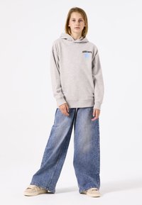 Sudadera de algodón gris con cordón, que presenta el texto "GOOD DAYS" y un gráfico colorido. Combinada con unos pantalones vaqueros de pernera ancha de denim azul y zapatos beige.