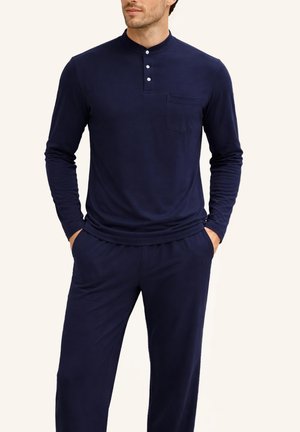 Mann trägt marineblaues Henley-Shirt mit langen Ärmeln und Knöpfen sowie passende Hose, Hände in den Taschen, vor einfachem hellem Hintergrund.