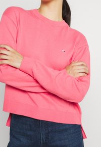 Rosa tröja i mjuk bomull med ribbad halsringning och nederkant, med en liten logotyp på bröstet och sidslitsar i botten.