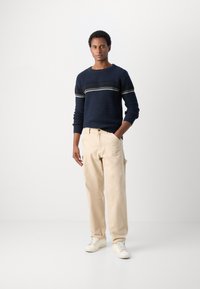 Marineblå strikkegenser med horisontale hvite og kremfargede striper, kombinert med beige bukser og hvite joggesko. Modellen står foran en nøytral bakgrunn.