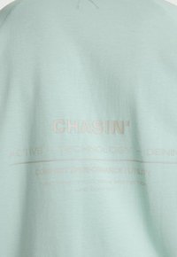 Tissu vert clair avec texte en relief "CHASIN'" et texte pâle en dessous indiquant "ACTIVE – TECHNOLOGY – DENIM" ainsi que des mots sur le confort et la performance.
