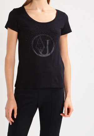 Kobieta nosząca czarną koszulkę z krótkim rękawem i okrągłym dekoltem, z ciemnym, okrągłym logo "AJ" i dekoracyjnymi kropkami na piersi.