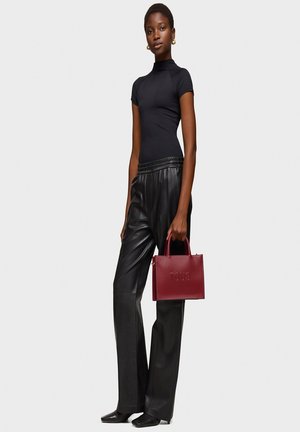 Parte superior negra de manga corta, pantalones de cuero negros holgados y un bolso de cuero rojo oscuro con el logo "TOUS" en relieve, sujetado en la mano derecha del modelo.
