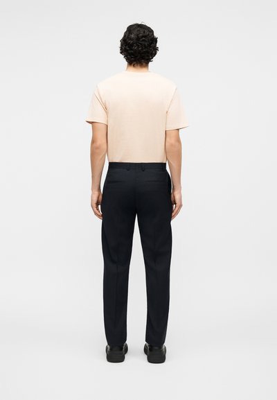 Άνδρας με σγουρά σκούρα μαλλιά στέκεται με την πλάτη, φοράει ανοιχτό ροζ κοντομάνικο πουκάμισο, μαύρο παντελόνι και μαύρα παπούτσια μπροστά σε απλό φόντο.