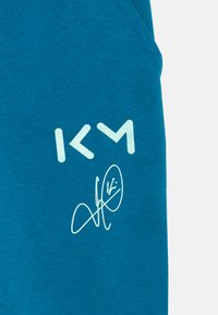 Teal sweatshirtstof med et hvidt stiliseret "KM" logo og et signaturdesign i lys blå, med en glat tekstur og uden synlige mønstre.