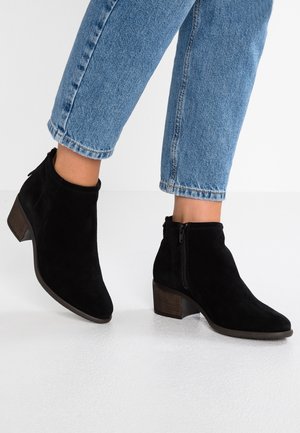 Jalkaterät mustissa mokkanahasta valmistetuissa nilkkabootseissa, joissa on lohko-otot ja sivuvetoketjut, yhdistetty vaaleansinisiin käärittyihin farkkuihin valkoisella lattialla.