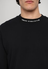 Felpa nera con colletto a costine. Presenta il testo bianco "VERSACE JEANS COUTURE" sul colletto. Texture morbida e liscia, design minimale.