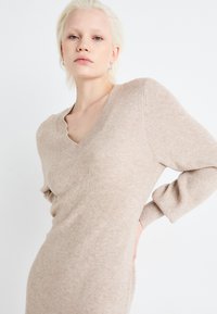 Abito lavorato a maglia beige con scollatura a V e maniche lunghe. Presenta un motivo textured e una silhouette aderente.