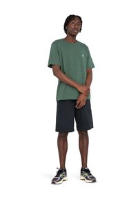 Grünes T-Shirt mit kurzen Ärmeln und Rundhalsausschnitt sowie kleinem Logo, kombiniert mit dunklen Shorts und gemusterten Sneakers. Insgesamt ein lässiges Outfit.