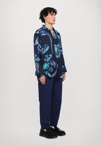 Marineblauw overhemd met kraag en lange mouwen, versierd met een turquoise bloemenpatroon, gedragen met donkerblauwe broek en zwarte schoenen.