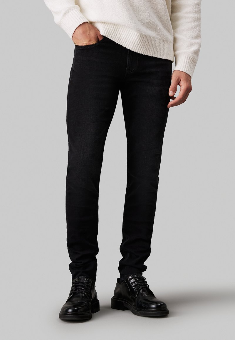 Jeans negros ajustados con una sutil textura, que cuentan con un diseño tradicional de cinco bolsillos y cierre de cremallera, combinados con botas negras de cordones.