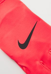 Gant de sport rouge en tissu extensible, doté d'un logo Nike en noir bien visible. Le design comprend des ouvertures pour les doigts et une texture lisse.
