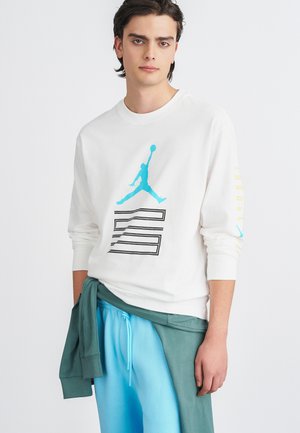 Valge pikkade varrukatega t-särk, millel on türkiissinine Jumpman logo ja must graafiline disain. Kanti koos helesiniste pükstega ja rohelise kapuutsiga särk, mis on üle heidetud.
