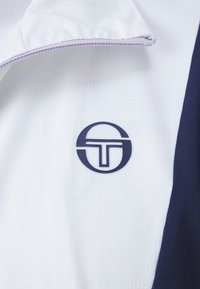 Reißverschlussjacke mit einem weißen und dunkelblauen Farbblockdesign, strukturiertem Stoff und einem kreisförmigen Logo, das prominent auf der Brust platziert ist.