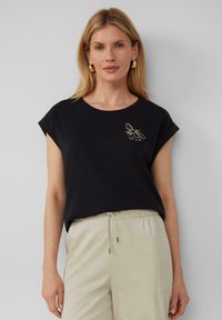 Zwarte t-shirt met korte mouwen en een ronde halslijn, voorzien van een gouden bij borduursel op de linkerborst. Gecombineerd met lichtbeige broek.