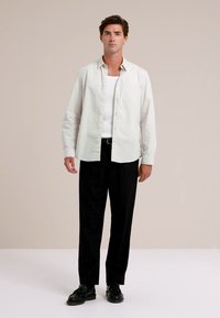 Jaunas vyras stovi vilkėdamas baltą atvirą marškinį virš baltos marškinėlių, juodas kelnes ir juodus batus prieš paprastą smėlio spalvos foną.