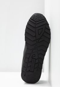 Semelle de chaussure en caoutchouc noir avec des crans profonds, inclinée contre un coin blanc, montrant le nom de la marque "ANNA FIELD" sur la semelle.