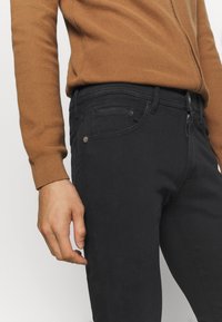 Närbild av en person som bär svarta tajta jeans och en brun texturerad långärmad tröja, stående med vänster hand avslappnad vid sidan.