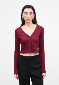 Cardigan cropped lung cu mânecă lungă de culoare burgundă, cu decolteu în V, având o serie de nasturi și textură ribbed, asociat cu pantaloni negri sobra.