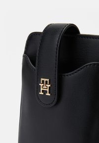 Geantă din piele neagră cu capac pliabil, având un accent logo de nuanță aurie și o suprafață texturată subtilă. Margini cusute vizibile.