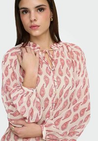 Blouse rose avec motif paisley rouge, encolure à volants, devant boutonné et manches avec poignets. Fabriquée en tissu léger à la texture douce.