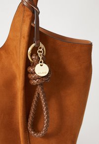 Borsa tote in suede marrone con tracolla in pelle intrecciata e accessori in oro. Presenta un'etichetta rotonda con la scritta "ALDO" in rilievo, che mette in evidenza il dettaglio di design.