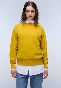 Sudadera amarilla con puños y dobladillo acanalados, que presenta un acabado texturizado. Llevada sobre una camisa blanca con cuello, combinada con jeans de color azul claro.