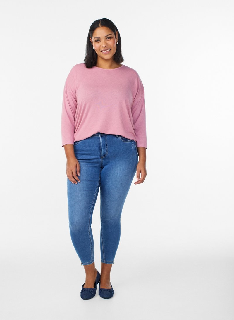 Femme souriante debout, portant un haut rose clair à manches longues, un jean skinny bleu, et des ballerines bleu marine sur fond blanc.