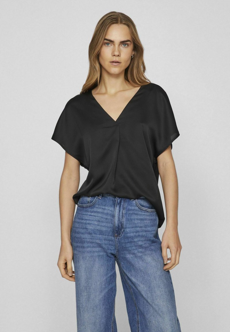 Top corto in satin nero con maniche corte, scollatura a V e piega frontale, abbinato a jeans in denim blu a vita alta. Texture semplice e liscia.