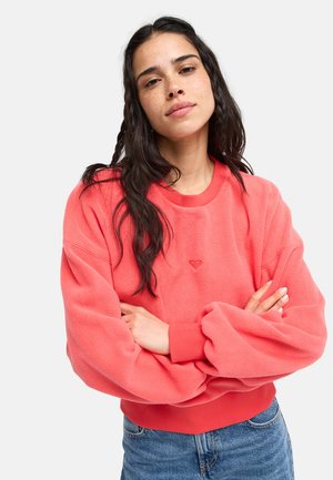 Femme aux longs cheveux foncés et taches de rousseur porte un sweatshirt corail avec les bras croisés et un jean bleu, debout devant un fond clair uni.