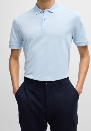 Homme portant un polo bleu clair à manches courtes avec le logo "BOSS", rentré dans un pantalon bleu marine foncé, mains dans les poches, vue de face.