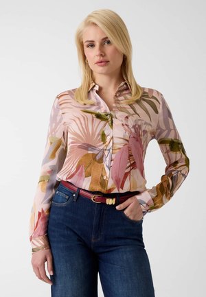 Blondynka w bluzce w kwiaty z długim rękawem, wsuniętej w ciemnoniebieskie dżinsy z czerwonym paskiem, stoi z jedną ręką w kieszeni.
