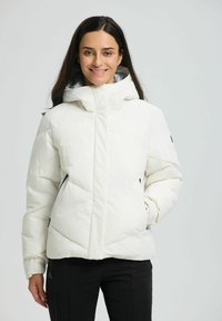 Smilende kvinde iført en hvid polstret vinterjakke med hætte, sorte bukser, der står imod en simpel lys baggrund.