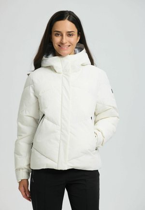 Glückliche vrouw die een witte gewatteerde winterjas met capuchon en zwarte broek draagt, staand tegen een effen lichte achtergrond.