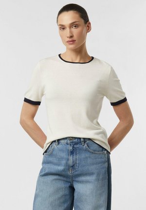 Kobieta o ciemnych włosach związanych do tyłu, ubrana w biały sweter z krótkim rękawem z czarnym obszyciem oraz niebieskie dżinsy z wysokim stanem, z rękami w kieszeniach.