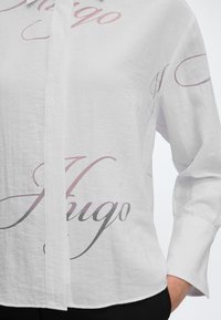 Chemise blanche boutonnée avec un grand texte cursif "Hugo" imprimé en rose clair et gris, portée avec une main dans la poche d’un pantalon noir.