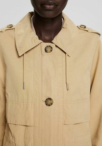 Gros plan d'une personne portant une veste beige avec de gros boutons en écaille de tortue et des cordons suspendus au col.