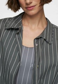 Donna che indossa una camicia a righe grigie sopra un top a righe grigie, con un primo piano del colletto e della parte superiore del torace.