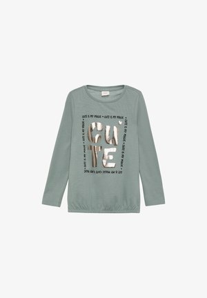 Langärmliges, helltürkisfarbenes Shirt mit gerafftem Saum. Mit metallisch silbernem Text, der "CUTE IS MY MAGIC" in einer verspielten Schriftart anzeigt.