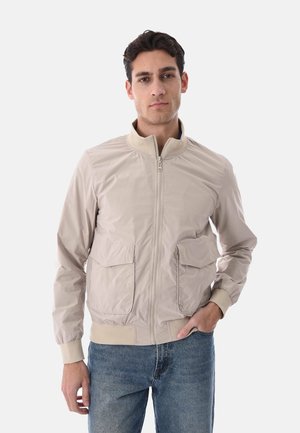 Uomo che indossa una giacca beige con zip e due tasche frontali con patta e jeans blu, in piedi su uno sfondo chiaro e uniforme.