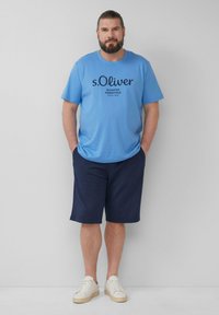 Lichtblauwe t-shirt met korte mouwen en zwarte opdruk, gecombineerd met marineblauwe shorts en witte sneakers, staande tegen een neutrale achtergrond.