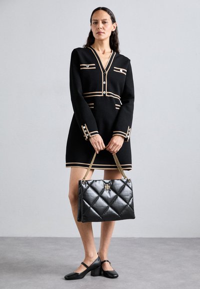 Vestido preto de malha com riscas douradas, mangas compridas e detalhes em botão. Segurando uma mala de mão preta almofadada com corrente dourada e detalhe de logótipo.