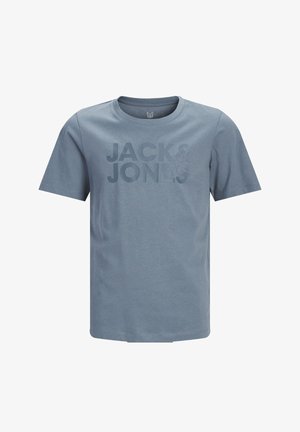 Young boy standing, wearing a blue "JACK & JONES" t-shirt, light wide-leg jeans, and beige sneakers on a plain white background.