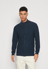 Suéter de cremallera azul marino con cuello alto, textura acanalada y mangas largas. Lleva puesto sobre pantalones de color claro frente a un fondo simple.