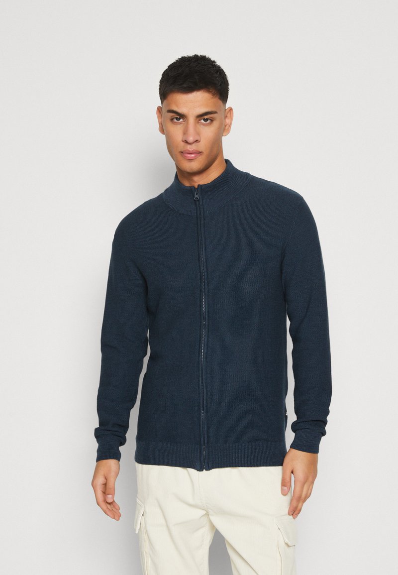 Granatowy sweter na zamek z wysokim kołnierzem, prążkowaną teksturą i długimi rękawami. Nosi go osoba w jasnych spodniach na jednolitym tle.