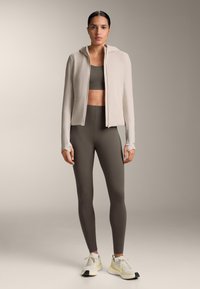 Ženska stoji v sivih pajkicah, ujemajočem športnem nedrčku, bež zadrgani jopiči z luknjami za palce in belih supergah z rumenimi poudarki na nevtralnem ozadju.