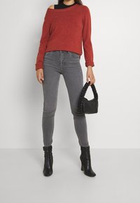 Nainen, joka pitää yllä ruosteenväristä olkapäältä avonaista neuletta, harmaita kapeita farkkuja ja mustia nilkkabootseja, sekä kantaa mustaa punoksista kahvaa olevaa käsilaukkua.