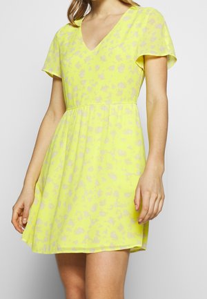 Kobieta ubrana w krótką, jaskrawożółtą sukienkę z delikatnym, jasnym kwiatowym wzorem i dekoltem w serek, stojąca na jednolitym tle.