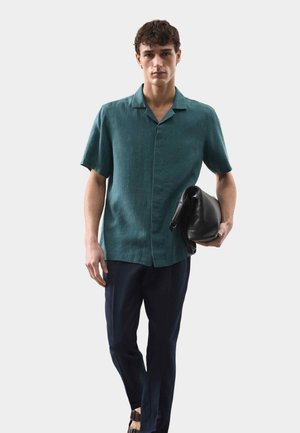 Mand iført en teal kortærmet skjorte med knapper og marineblå bukser, holder en sort foldet lædertaske mod en ensartet lys baggrund.