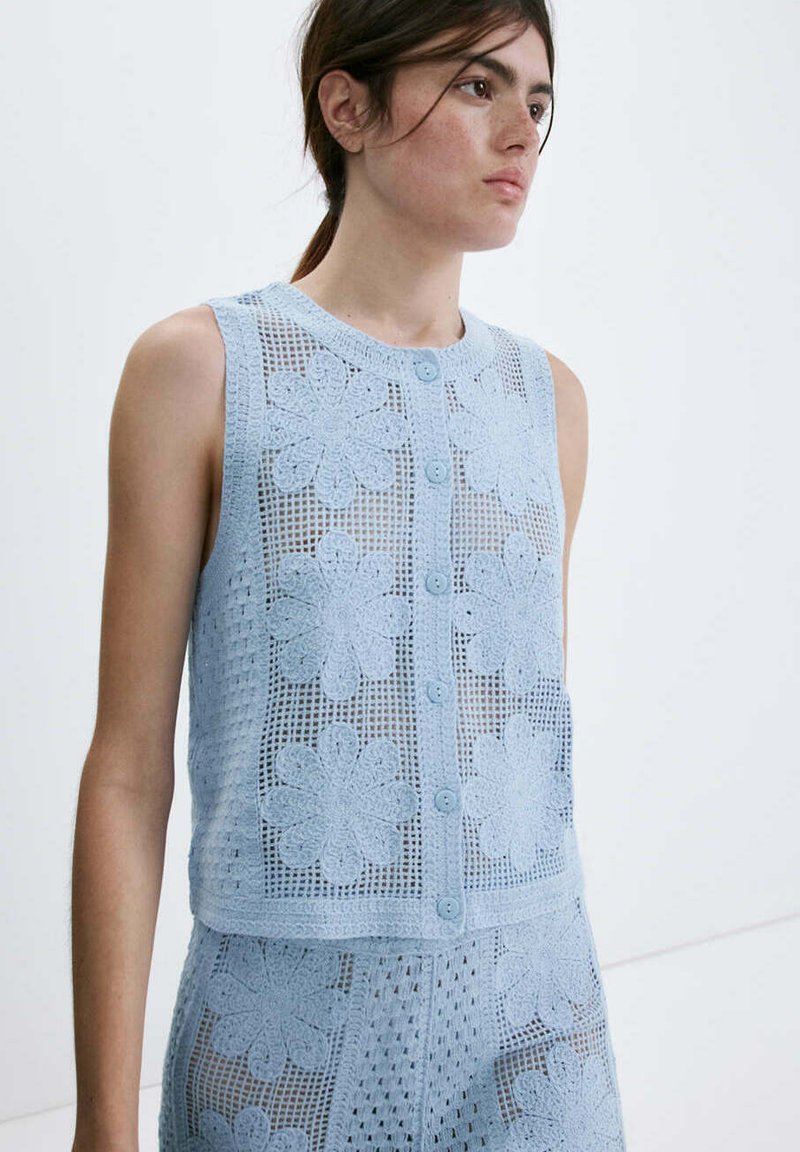 Jeune femme portant un haut sans manches bleu clair en crochet fleuri et un short assorti, debout devant un fond blanc uni.